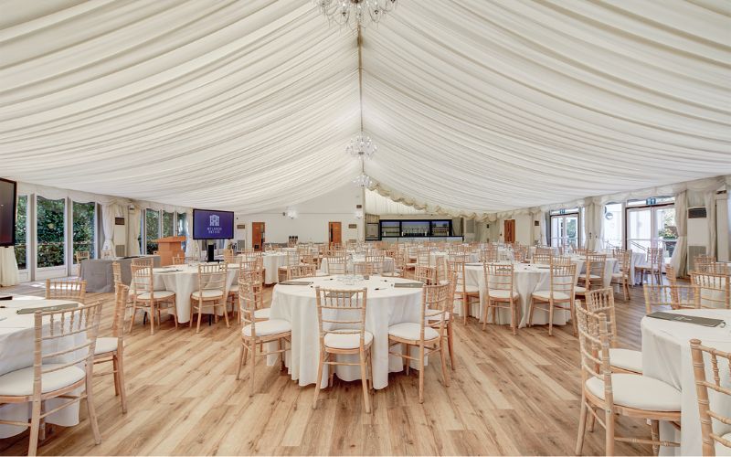 The Grand Pavilion set up with round tables and chairs. There is a podium up front and a large TV.