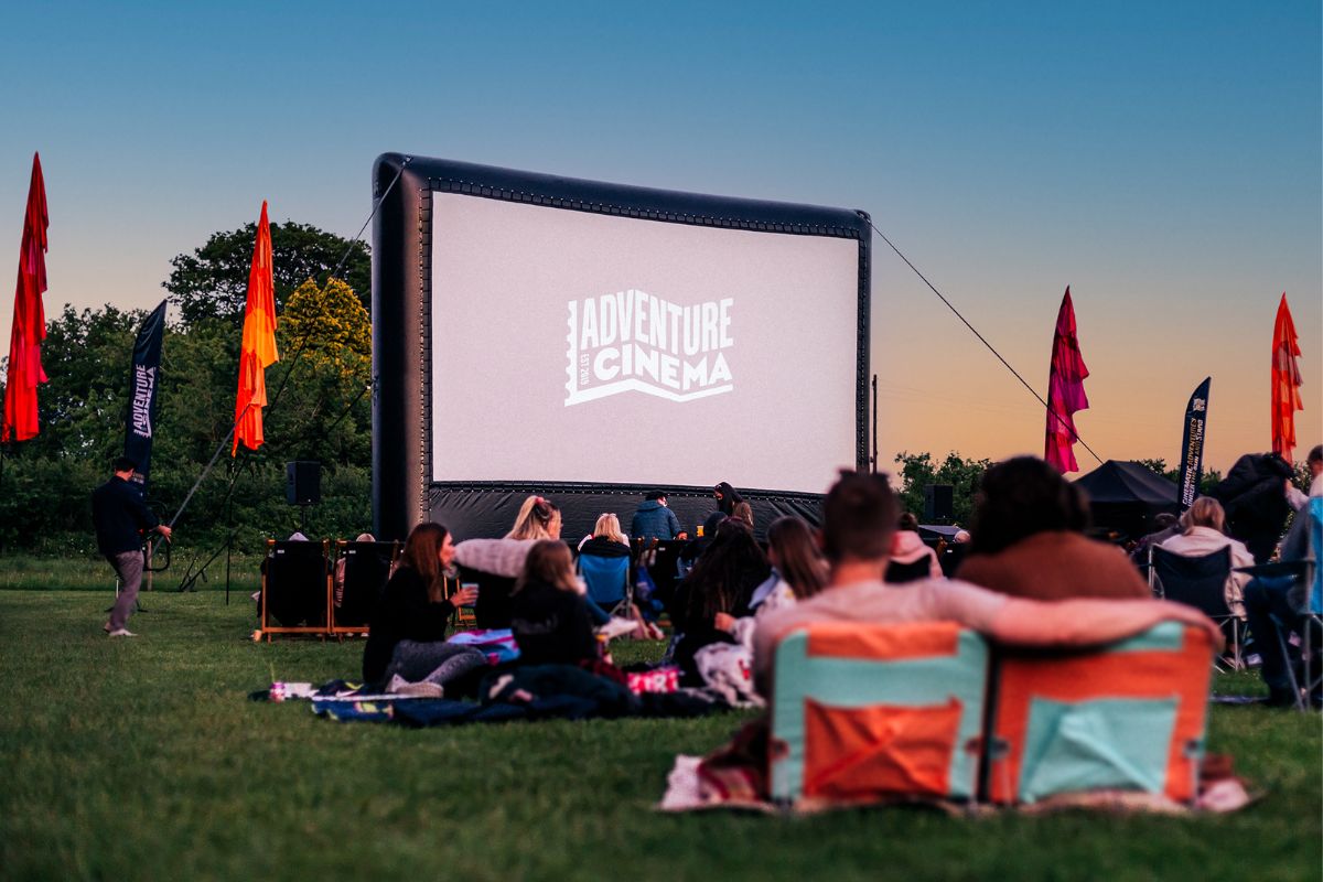 The Adventure Cinema outdoor screen with people sat on the grass in front.